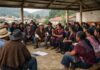 Sur-sureste de México: ¿persecución religiosa o choque de culturas? Personas indígenas, hombres y mujeres de distintas edades, sentadas en círculo bajo un techo comunitario, participando en una asamblea; algunos hablan mientras otros escuchan, con casas y montañas al fondo.