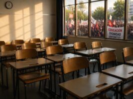 La CNTE y los límites de una movilización Salón de clases vacío iluminado por la luz de la tarde, mientras al fondo, detrás de las ventanas, se observa una movilización social desenfocada. La imagen simboliza la tensión entre el derecho a la protesta y el impacto de los conflictos laborales en la educación de los estudiantes.