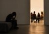 ¿Son los pobres más religiosos? Persona sentada en soledad y en penumbra mientras, al fondo, un pequeño grupo conversa reunido en un espacio iluminado, como representación de la necesidad, la búsqueda y el encuentro con una comunidad.